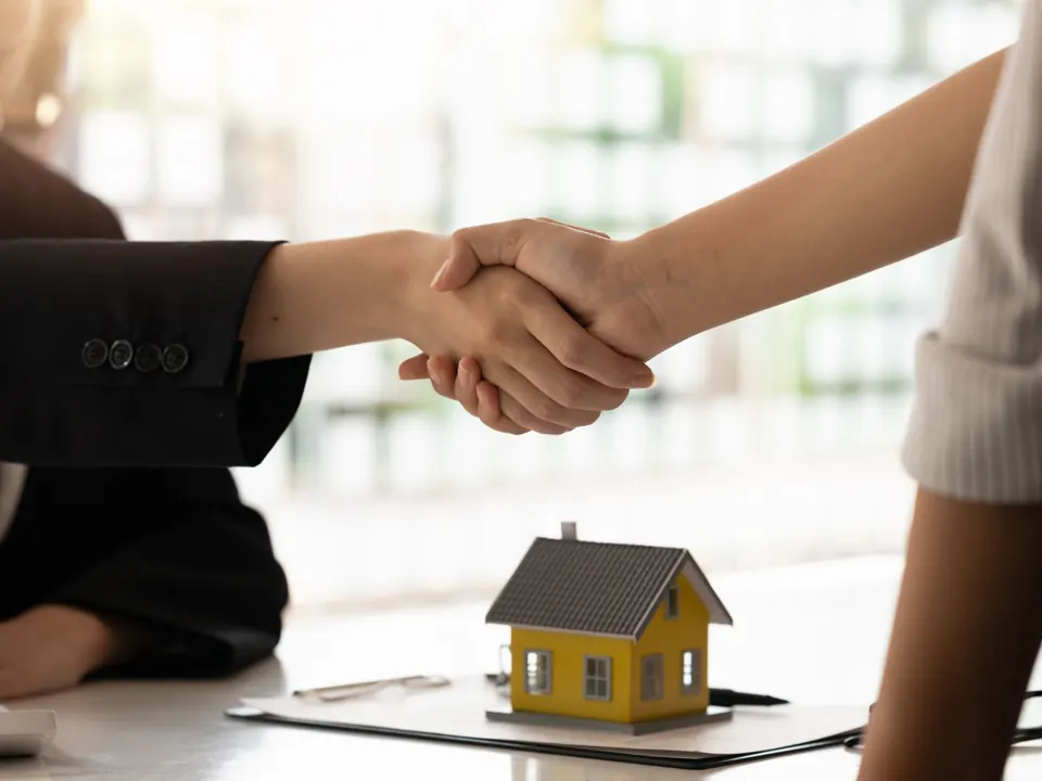 Aperto de mãos fechando negócio, com maquete de casa sobre a mesa